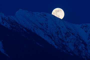 The full moon rises above snow covered mountain peaks on a spring night in the Coast Mountains of British Columbia.