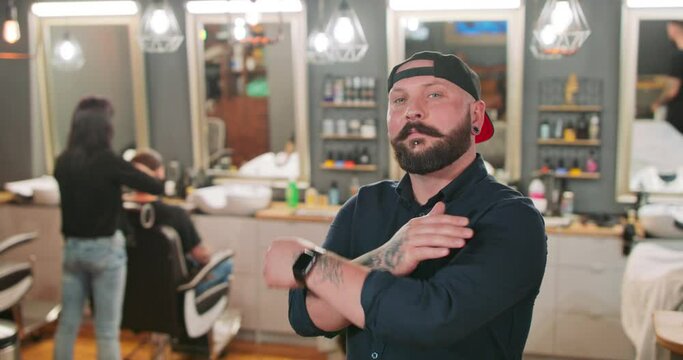 Young bearded and mustashed man, with tattoed hands, with chin ring and tunnel earrings, in baseball hat, in front of camera, crosses his arms over his chest in barbershop. In the background the