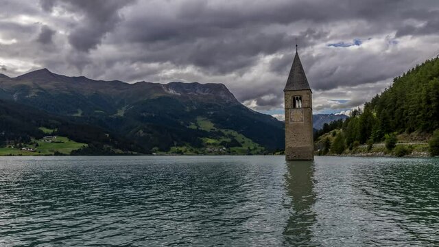 The top of the old bell tower of Curon (Graun) emerges from the waters of Lake Resia, against a dramatic sky, South Tyrol, Italy. Time lapse motion