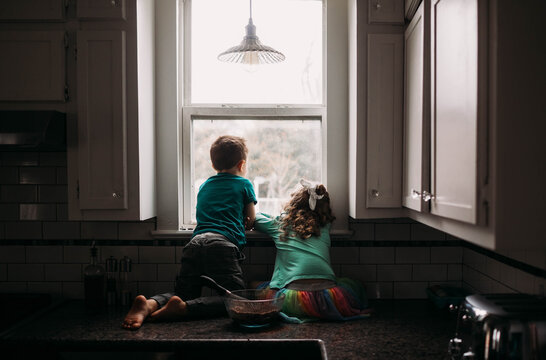 Rear View Of Brother And Sister Looking Through Kitchen Window During Rain