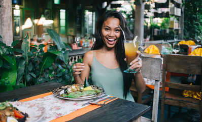 Happy young ethnic lady enjoying cold natural juice and exotic food in cafe