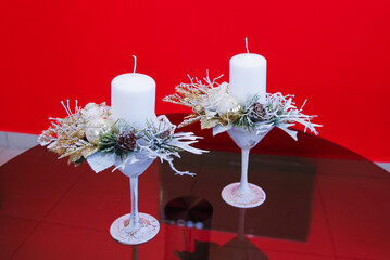 Winter compositions in snowy champagne glasses made of white candles, white and gold lights, glass balls, and cones stand on a quilted table against a red background. NEW YEAR home interior