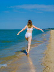 Fototapeta na wymiar Rear view of a girl running away into the distance along a sandy sea beach Splashes of sea water. The girl is wearing a white swimsuit and sunglasses. Freedom. Fitness. Summer sunny day. Copy space