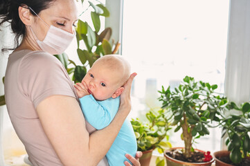 Beautiful mother in protective face mask holding her little cute newborn son protecting him from viruses and infections. Coronavirus covid-19 prevention. Home quarantine. Family at home by the window.