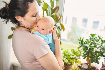 Beautiful mother in protective face mask holding her little cute newborn son protecting him from viruses and infections. Coronavirus covid-19 prevention. Home quarantine. Family at home by the window.