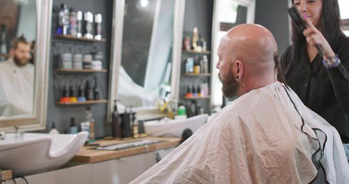 A female barber turned to camera, brushes bearded man's smoothly shaved head without hair, who is sitting on the barber's chair, facing the mirror, and talking. Grey wall with mirrors and cosmetics on