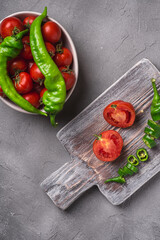 Fresh ripe tomatoes in bowl near to old wooden cutting board with hot chili peppers, stone concrete background, top view