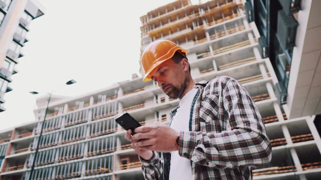 Caucasian male construction worker in protective helmet use smartphone near building that is being built. Engineer checks construction process, makes notes on phone. Foreman chatting in application