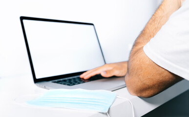 Person working from home at their desk with a laptop and a medical face mask on top of a white desk.