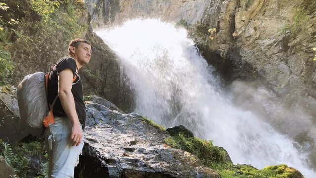 Portrait of young man looking away on background of waterfall. Adult male tourist enjoying beautiful view in mountainous area.