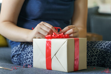 Close up of unknown caucasian woman midsection and hands sitting on sofa at home with making or opening wrapped box with red tie ribbon - birthday or holiday present giving and receiving concept