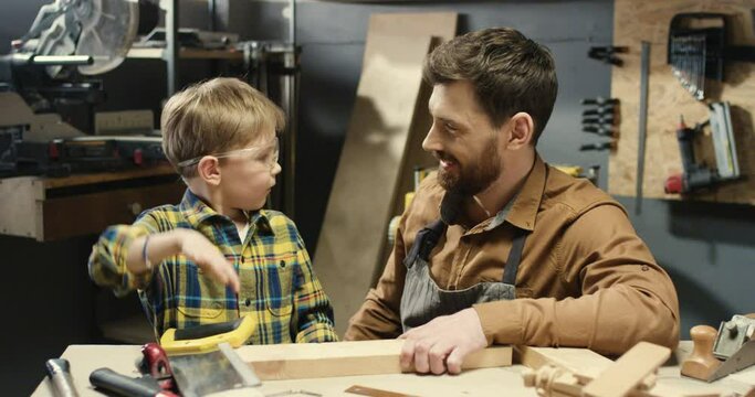 Caucasian woodman happy father teaching his little son to work with hardwood and sawing timber. Handsome man dad carpenter showing to small boy filing wood in workshop.