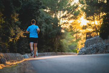 Young man wearing blue shirt and black shorts runs on an idyllic road during sunset surrounded by nature in Mallorca (Spain)