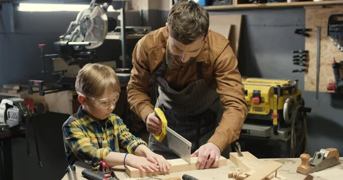 Caucasian woodman happy father in goggles teaching his little son in glasses to work with hardwood and sawing timber. Handsome man dad carpenter showing to small boy filing wood in workshop.