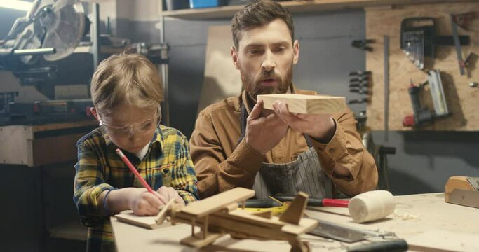 Cheerful handsome Caucasian young father with little cute son handcrafting in workshop with wood. Dad teaching small boy to work with timbers. Carving working concept of two generations.