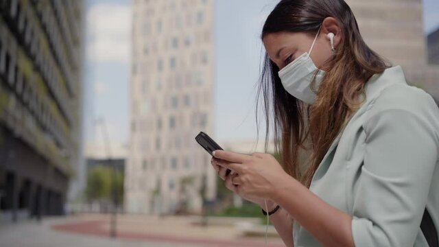 Close-up of a working woman in a face mask using a smartphone with headphones.