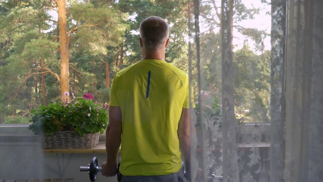 A man in sportswear playing sports with dumbbells on the balcony of his apartment during quarantine and self-isolation