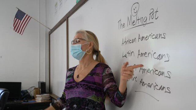 Camera Pushes In On Female Civics Or Grammar Teacher Wearing Medical Face Mask, Writing America The Melting Pot On A White Board In A School Classroom. Concept Posing Question For Students.