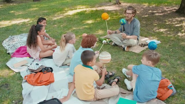 Medium shot of little cheerful schoolchildren and bearded male science teacher sitting on blankets in park on summer day and holding models of planets while learning about solar system