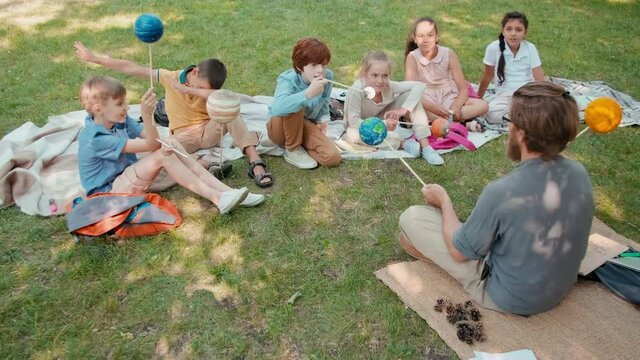 Tracking shot of male teacher with beard and glasses and group of schoolchildren sitting on blankets in park and holding models of planets while learning outdoors