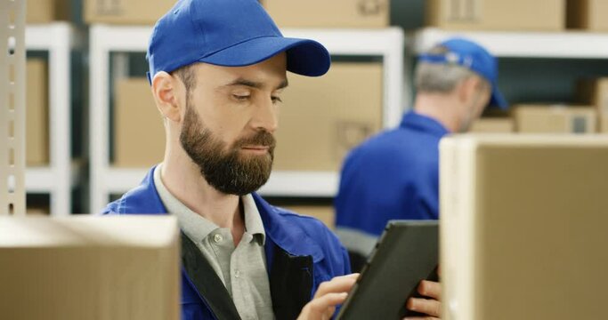 Close up of handsome Caucasian postman in blue uniform and cap standing between shelves with carton boxes in postal office storage. Man with tablet device searching for parcel and registering mails.
