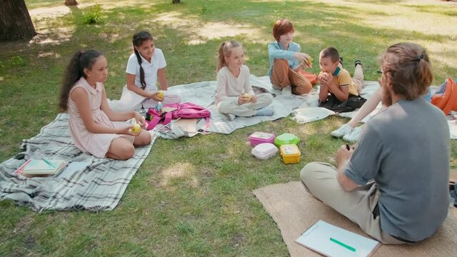 Tracking slowmo shot of male teacher with magnifying glass sitting on blanket in park and talking to group of schoolchildren listening to him and eating lunch after outdoor lesson