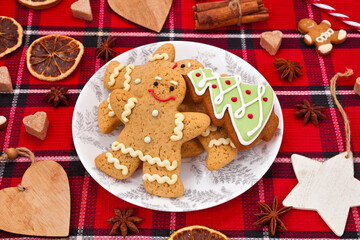 Christmas greeting card. Gingerbread men with fir shaped cookie in a plate, sugar hearts, dried orange slices, red berries and spices on a red fabric background.