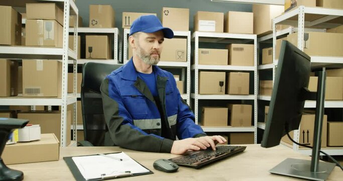 Handsome Caucasian postman in iniform and cap sitting at table in postal office store and working at computer. Male courier typing on keyboard in storage of parcels. Man browsing online.