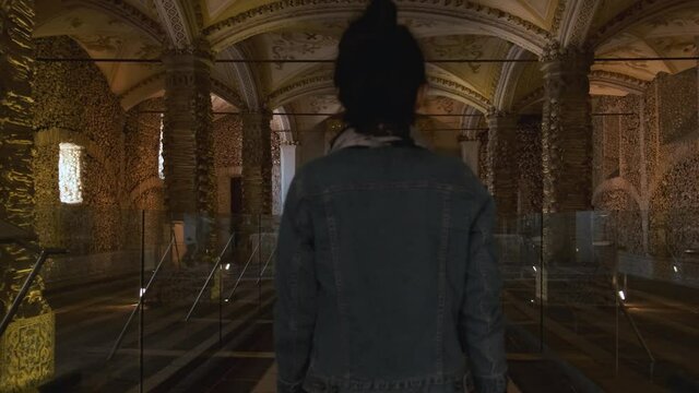 Adult woman walks inside Evora chapel of bones (Capela dos Ossos) on walls and pillars decorated, arranged and covered with human skulls and remains in cement, Portugal, static close up behind slowmo