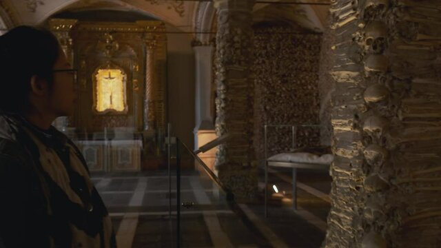 Adult Asian woman walks inside Evora chapel of bones (Capela dos Ossos) and looks at walls decorated and covered with human skulls and bones in cement, Portugal, static close up profile slow motion
