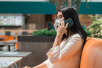 woman with black hair sitting while talking on the phone, wearing a mask, glasses and a white blouse, has a table in front