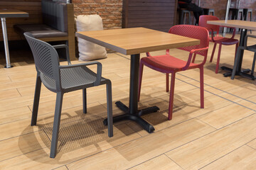 small wooden and metal table with two chairs, one red and the other gray, with a wooden floor, in the background there are other tables, chairs and a sack