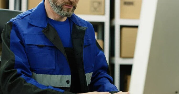 Close up of Caucasian postman in iniform sitting at table in postal office store and working at computer. Man typing on laptop in mail storage of boxes.