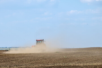 Fototapeta premium A red farm tractor in a cloud of dust cultivates the soil in the field with a cultivator after harvest. Summer sunny day. Fertile land. Modern agricultural machinery. Copy space. High quality photo