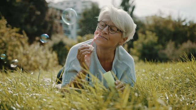 Retired senior woman enjoying freedom. Childish grandma blowing soap bubbles in the park. High quality 4k footage