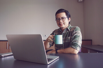 Asian man sitting with a laptop drinking coffee at his desk