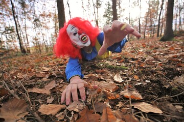 Halloween holiday. A scary evil clown crawls through the autumn foliage and stretches out his hands.Halloween carnival party outdoors.Masquerades and festive carnivals in October.Horror and fear.