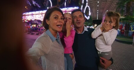 A happy carefree smiling family is having fun to make selfie or video call to friends or relatives while enjoying time together in amusement park with luna park lights at night.