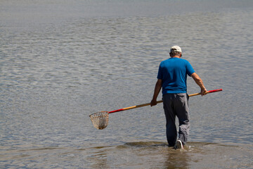 An elderly caucasian man wearing water resistant pants and baseball hat is walking in shallow water...