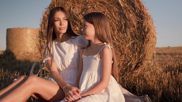 Mother and daughter in a white dress sitting in a field. Mom kisses her daughter on the forehead and hugs her