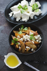 Black bowl with baked potato wedges served with feta cheese in a greek style, vertical shot on a brown stone surface