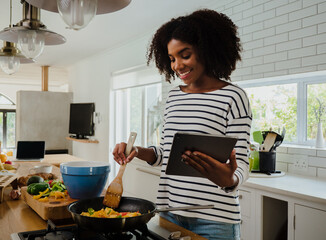 Smiling mixed race female chef reading online recipe holding digital tablet making hot curry in modern kitchen.