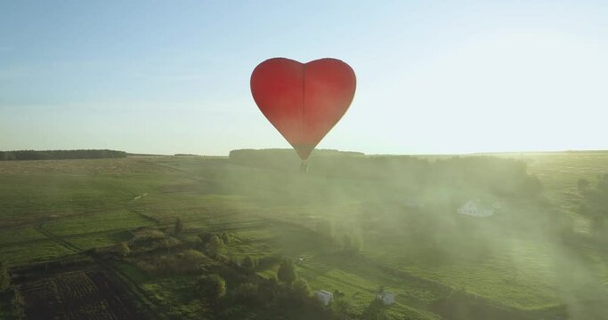 Red hot air balloon shaped heart flight above the village with smoke from chimney at summer sunny day - Aerial drone view