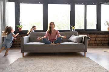Calm woman concentrating on yoga exercises on couch at home while two noisy kids laughing, running,...