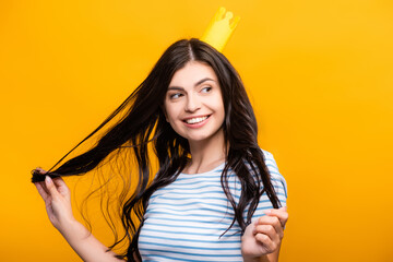 brunette woman in paper crown touching hair and smiling isolated on yellow