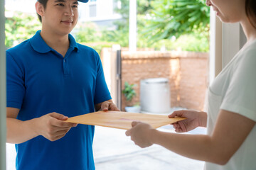Asian delivery young man in blue uniform smile and holding letter or document envelope in front house and Asian woman accepting a delivery of envelope from deliveryman. Advertising, Transportation.