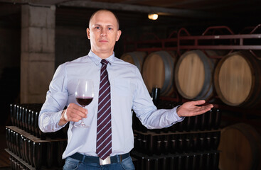 Glad male winemaker giving tour around winery, inviting to tasting red wine of own production