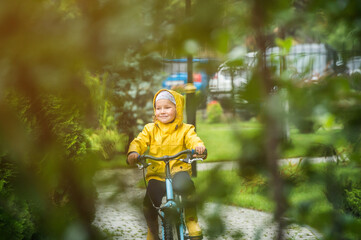 Happy little kid girl riding on bicycle in park. Child in waterproof yellow coat and rubber boots. Kid having fun on rainy day on bike. Playing on bike on sunny autumn rain day.