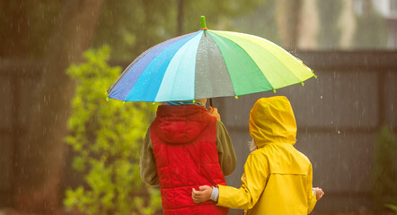 Two adorable children, boy and girl playing in park with colorful rainbow umbrella on a rainy autumn day. Child in waterproof yellow coat and rubber boots. Kid having fun on rainy day