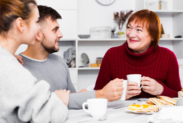 Senior woman with young couple enjoying time at home, drinking tea with sweets